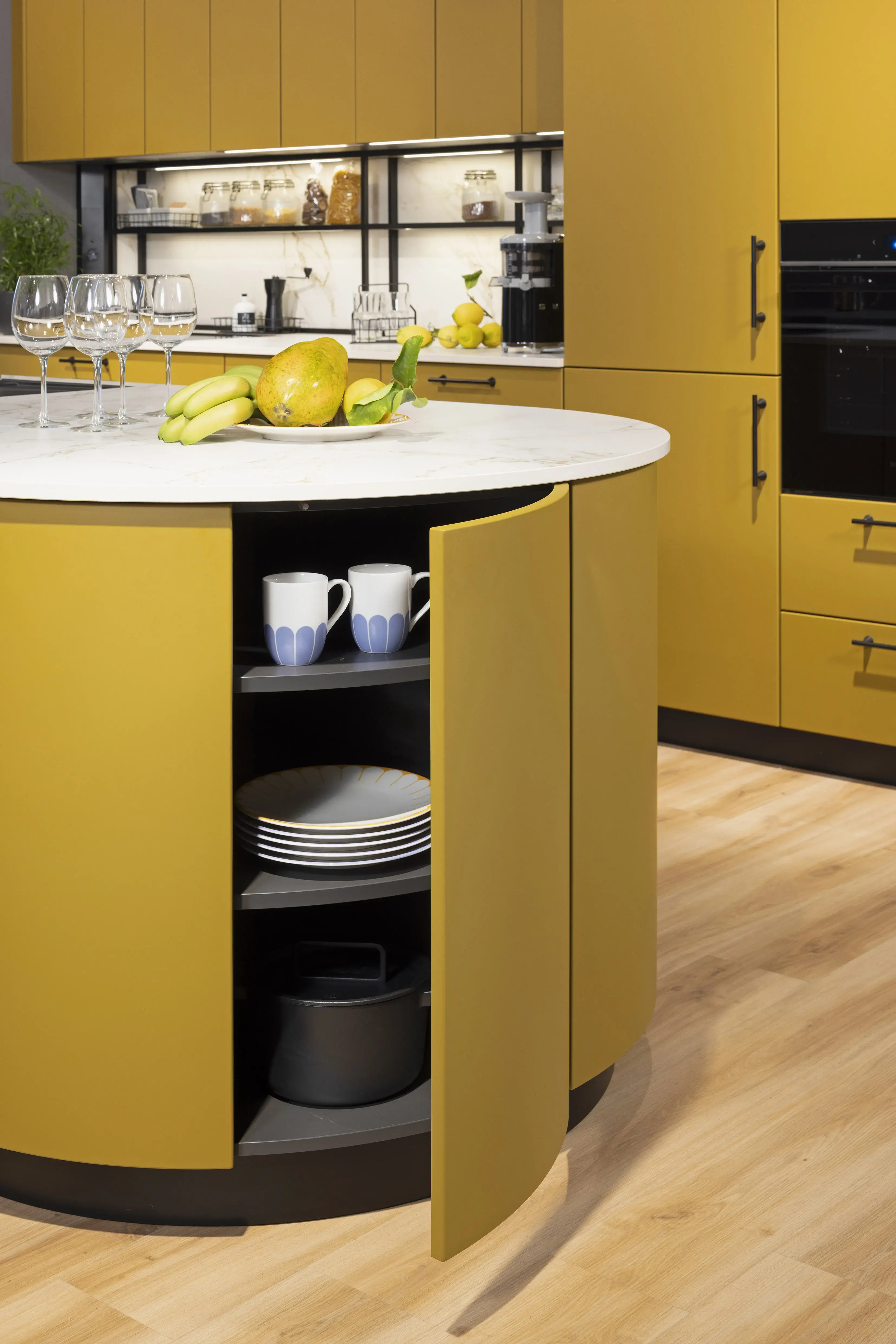 Moderne gelbe Küche mit runder Insel, offenem Schrank und Obst auf der Arbeitsplatte.