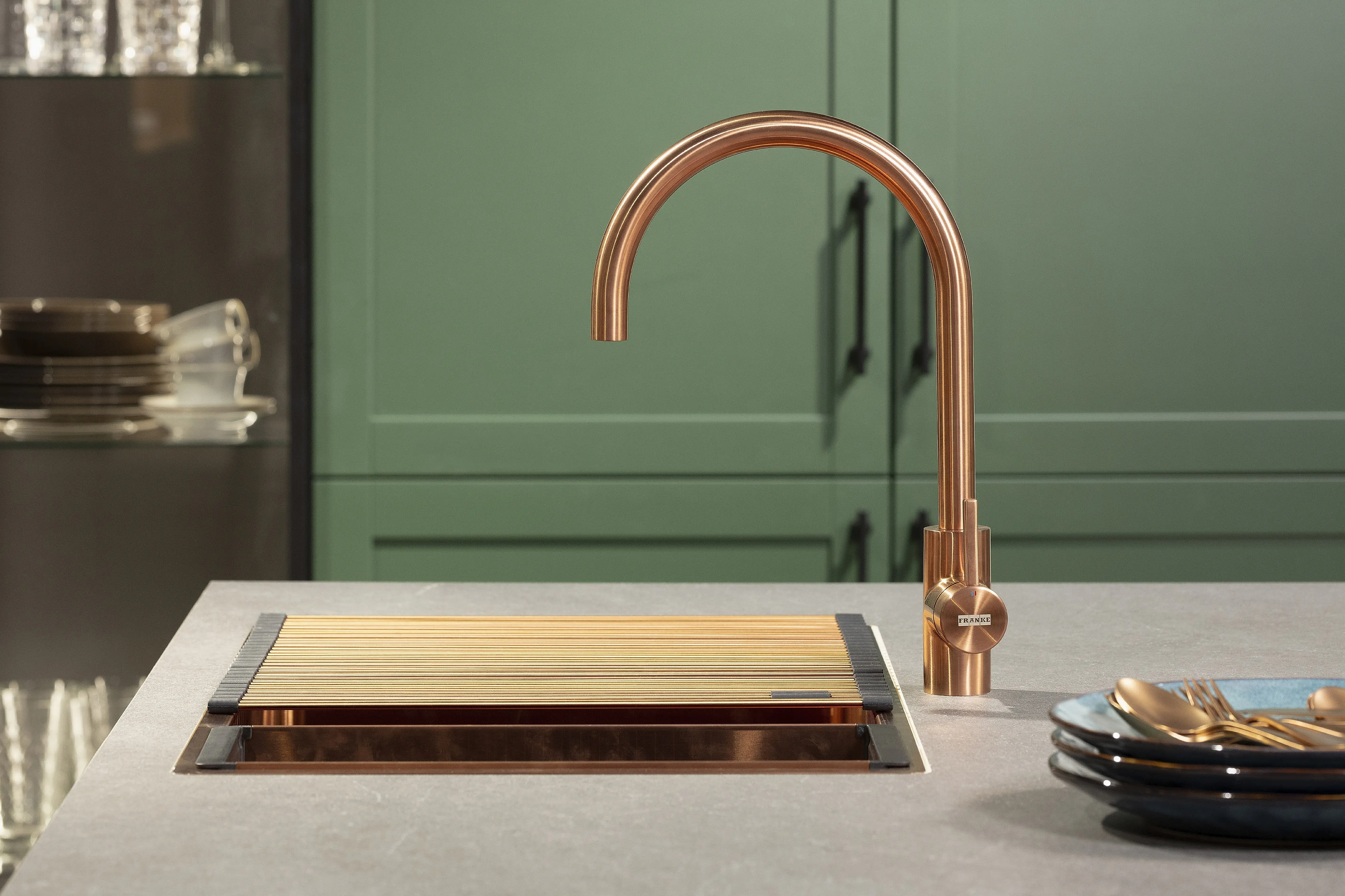 Kupferfarbener Wasserhahn an moderner Küchenspüle mit Holzbrett und grünen Schränken.