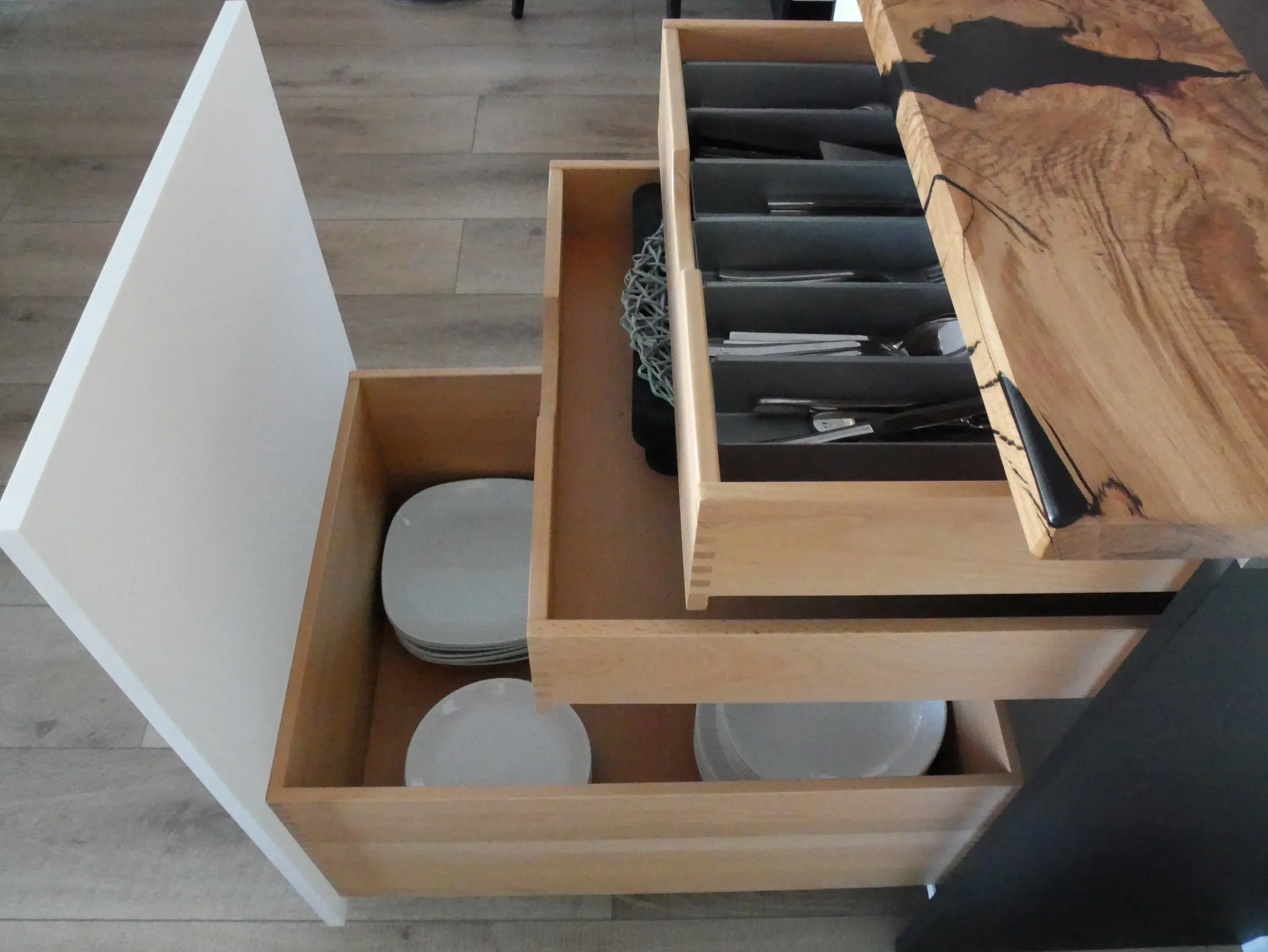 Offene Küchenschubladen mit Besteck und weißen Tellern auf Holzfußboden.